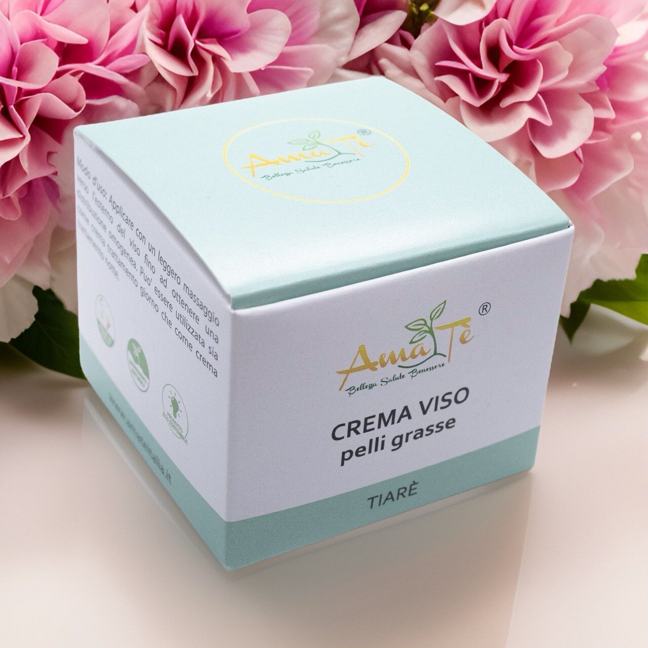 Confezione di Amatè Crema Viso pelli grasse Tiarè davanti a fiori. Scatola bianca con dettagli verde chiaro. Nome e logo visibili.
