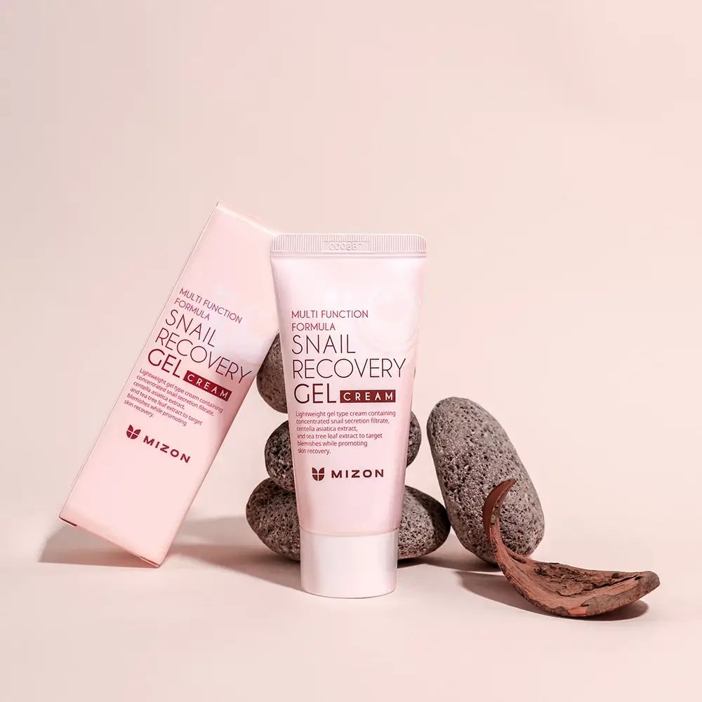 Tubo rosa con tappo bianco e scatola. Scritta: Snail Recovery Gel Cream. Marchio: MIZON. Appoggiato su pietre.