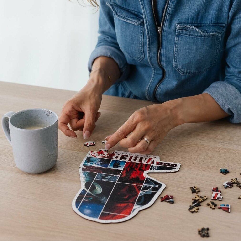 Persona che assembla un puzzle di Darth Vader. Puzzle in legno con motivi di Star Wars. Pezzi su un tavolo. Tazza di caffè accanto.