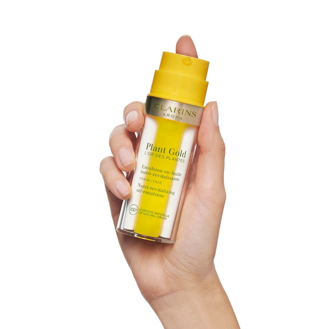 Mano che tiene Clarins Plant Gold emulsione olio. Flacone giallo con pompa, base trasparente. Testo: Emulsion-en-huile, Nutri-revitalisante.