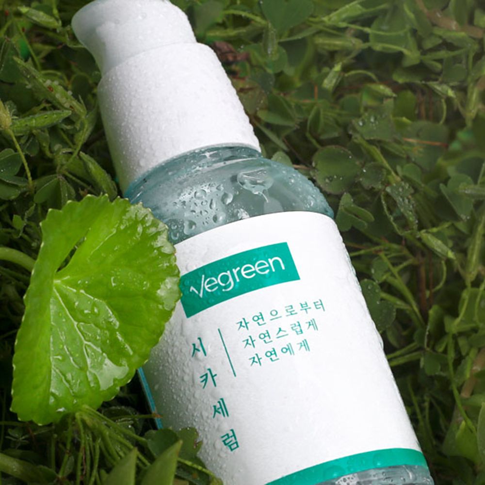 Flacone di siero per il viso nell'erba. Tappo a pompa bianco, etichetta verde con testo. Gocce sul flacone e una foglia.