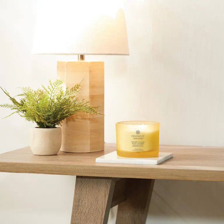 Candela gialla su un tavolo, accanto a una lampada e una pianta in vaso. Scritta: 'Chesapeake Bay Mind & Body'.