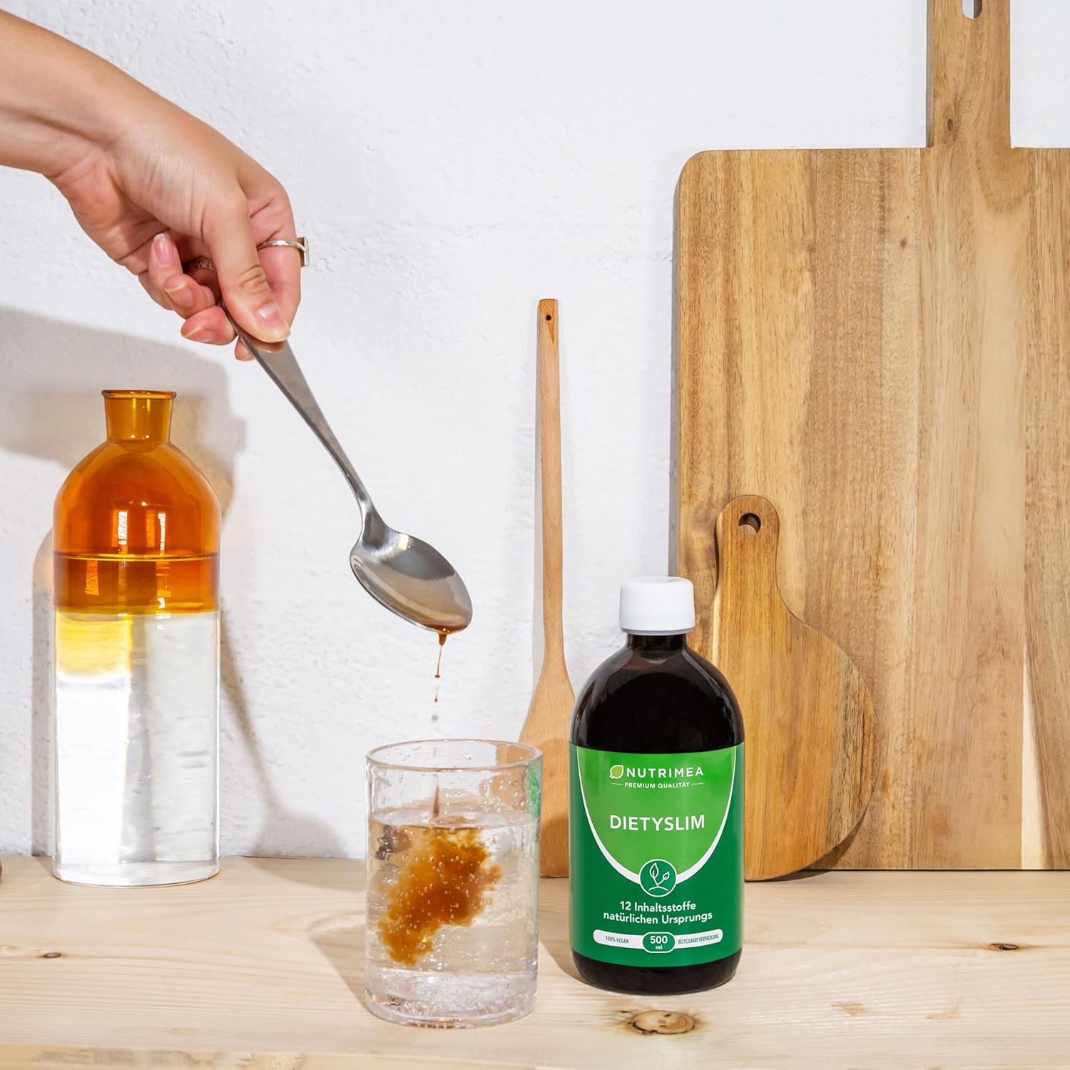 Una mano versa liquido da un cucchiaio in un bicchiere. Accanto, una bottiglia con etichetta verde.