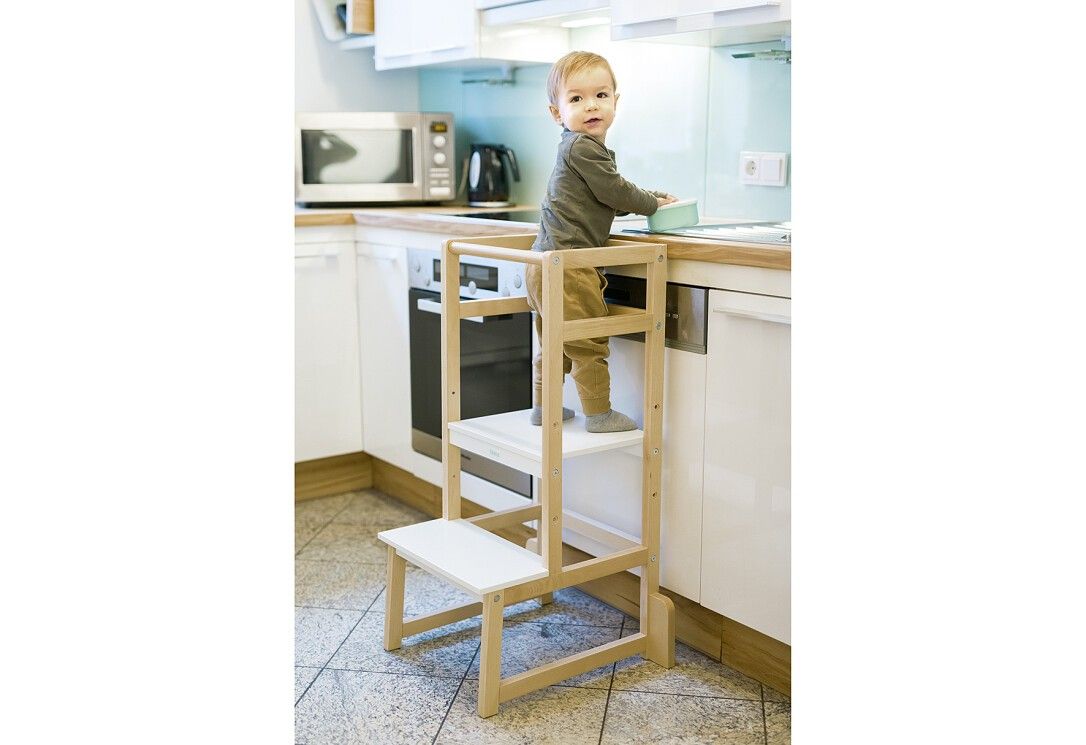 Bambino in piedi su una torre di apprendimento in una cucina. Struttura in legno con gradini bianchi. Il bambino afferra un oggetto sul piano di lavoro.