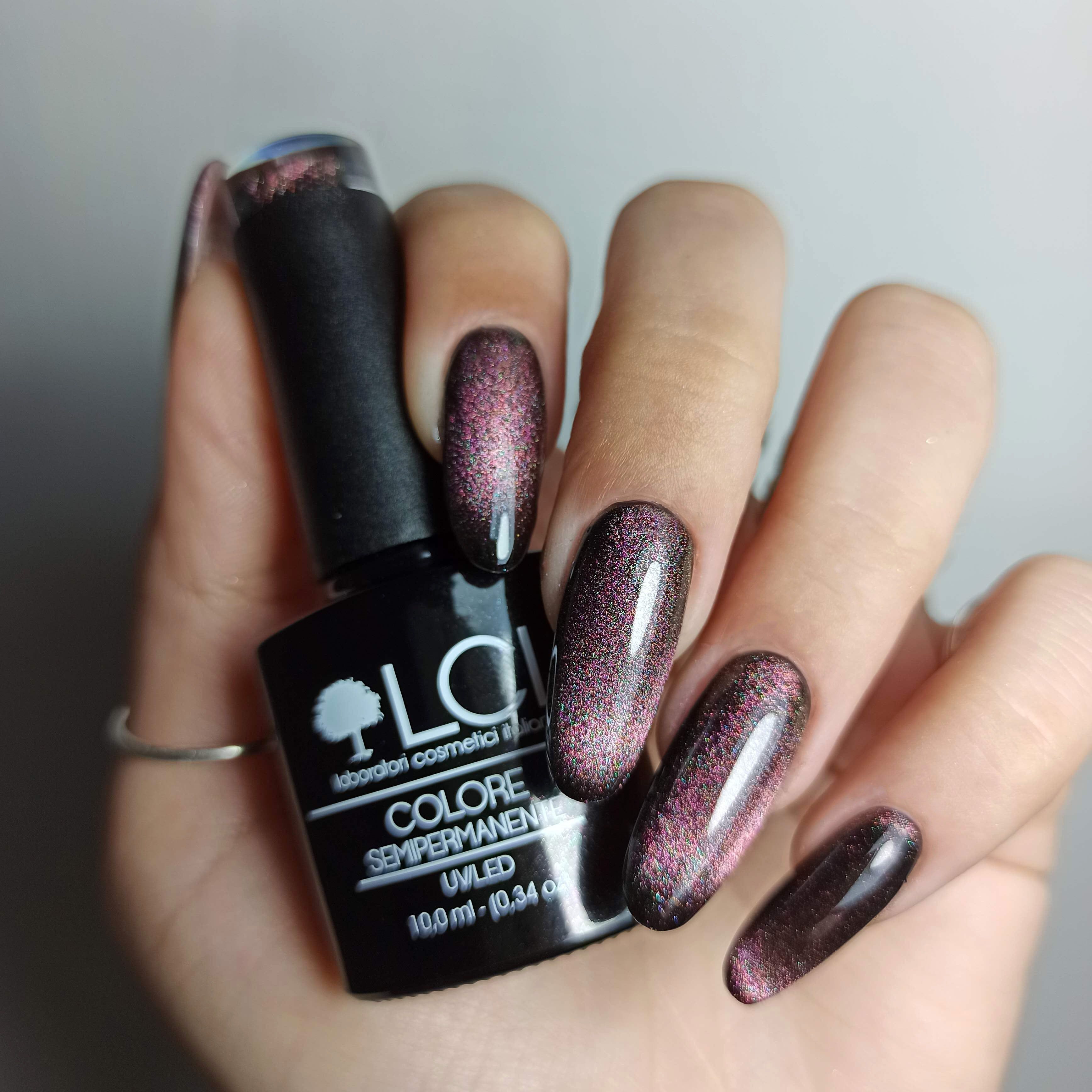 Mano con unghie smaltate e flacone di smalto. Unghie con effetto occhio di gatto rosa-marrone. Scritta: LCI, COLORE SEMIPERMANENTE, UVLED 10,0 ml.