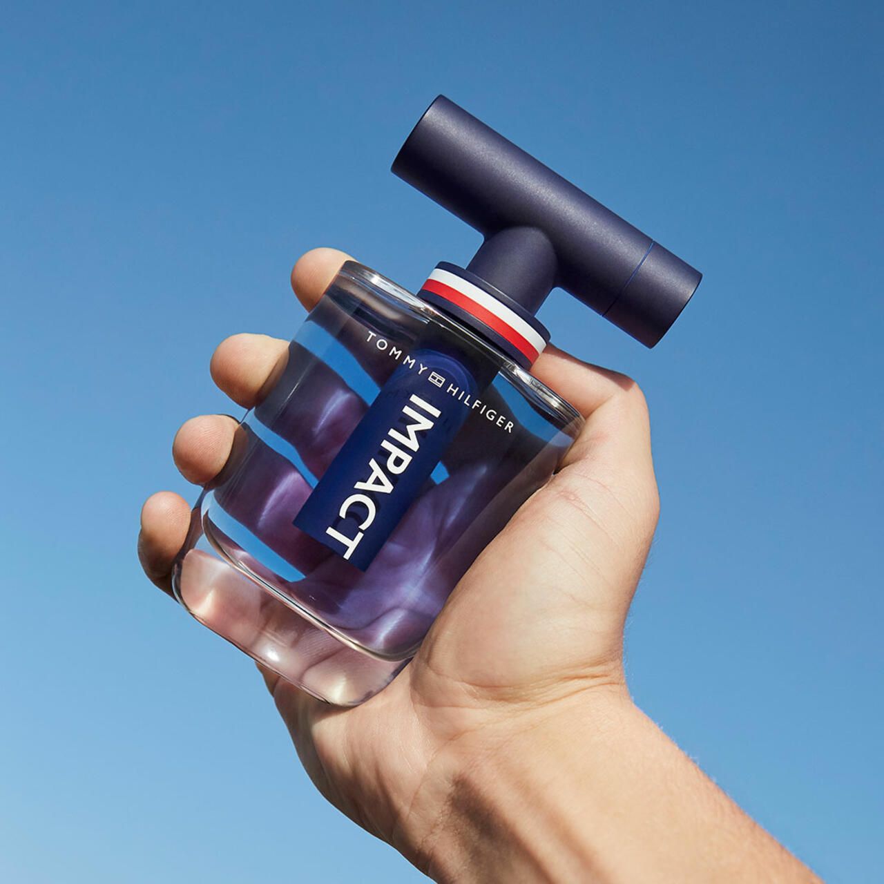 Mano che tiene un flacone. Flacone con contenuto blu e tappo nero. Scritta IMPACT e Tommy Hilfiger.