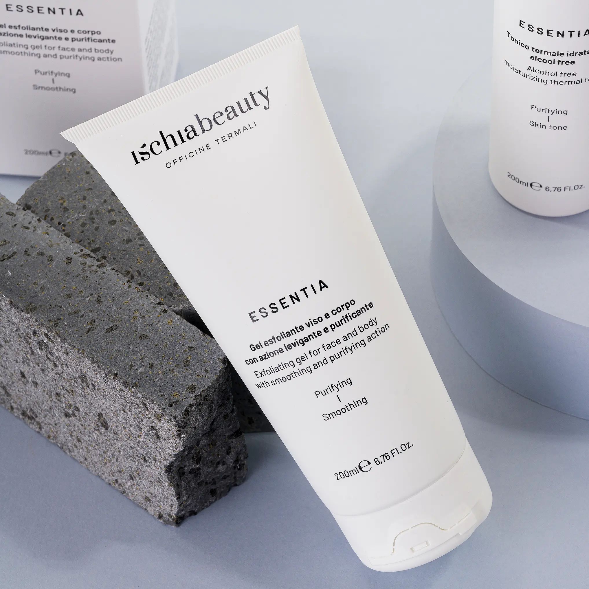 Tubo bianco con testo del prodotto. Testo: Ischiabeauty, Essentia, Gel esfoliante viso e corpo. Posizionato su un blocco di pietra, accanto a un altro tubo.
