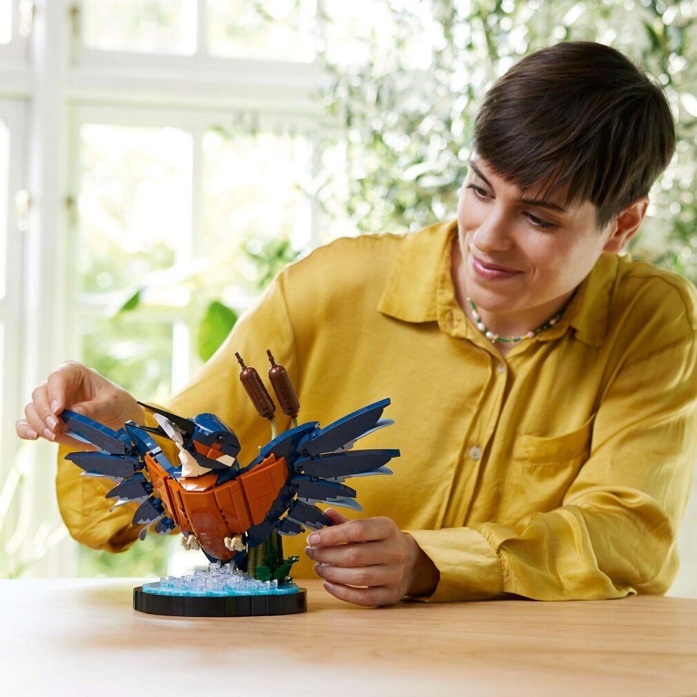 Persona che costruisce LEGO Icons 10331 Martin Pescatore. Modello blu-arancio con ali spiegate, su base con canne. Su tavolo.
