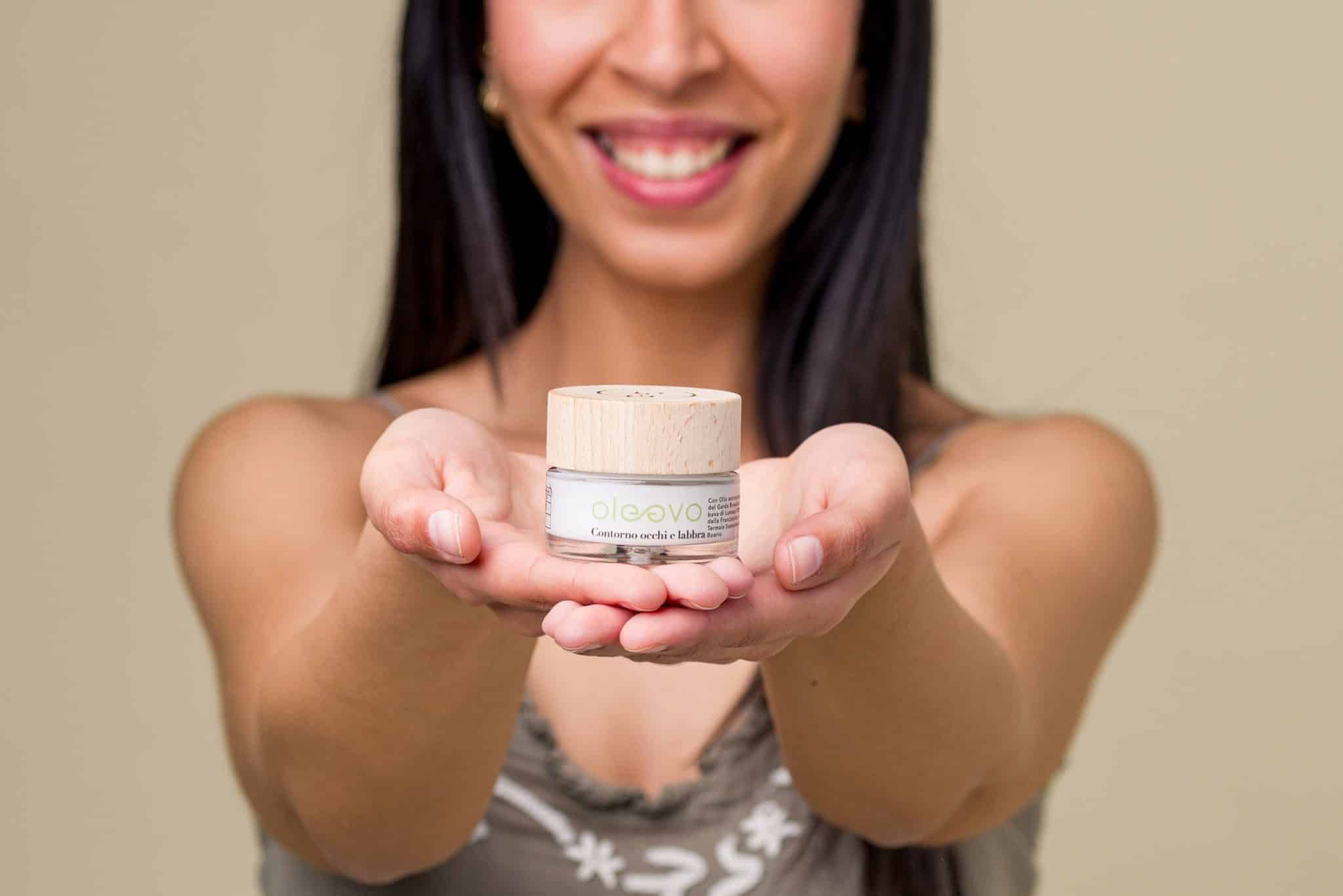 Donna che tiene un prodotto tra le mani. Vasetto di vetro con coperchio in legno. Scritta: Oleevo Contorno occhi e labbra.