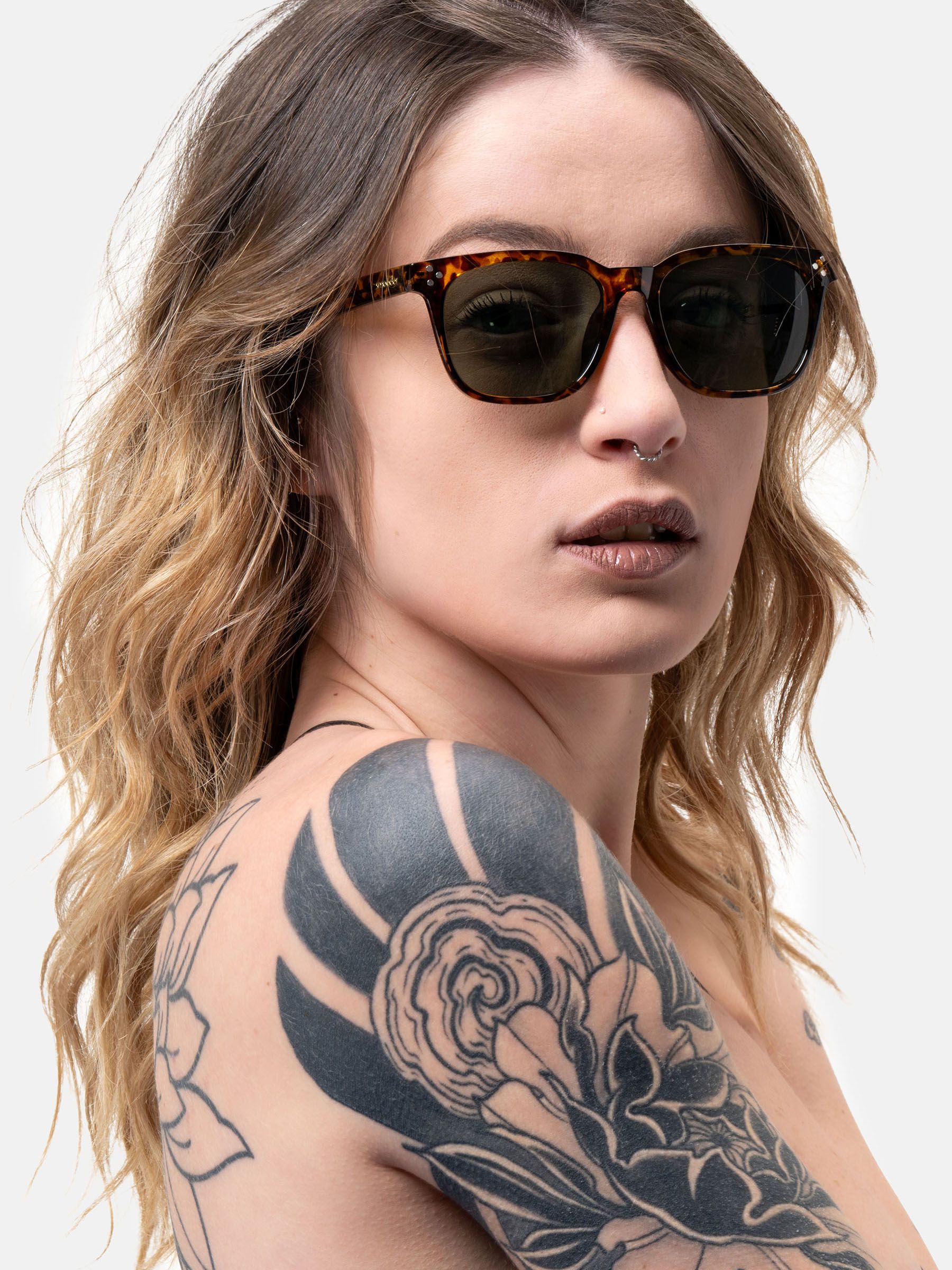 Persona che indossa occhiali da sole marrone tartarugato e lenti verdi. Forma quadrata. Borchie sulle aste.