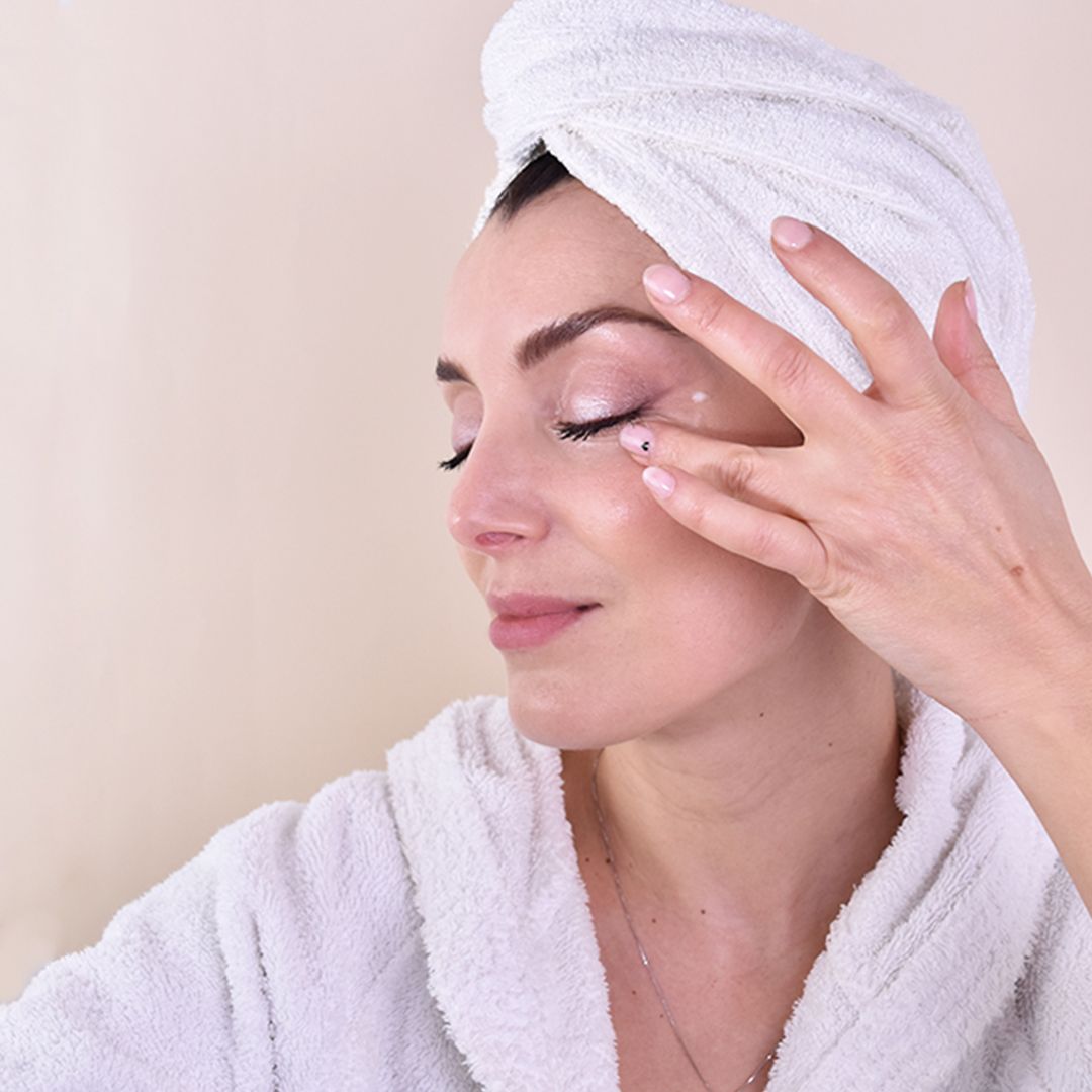 Donna con asciugamano in testa applica crema sotto gli occhi.