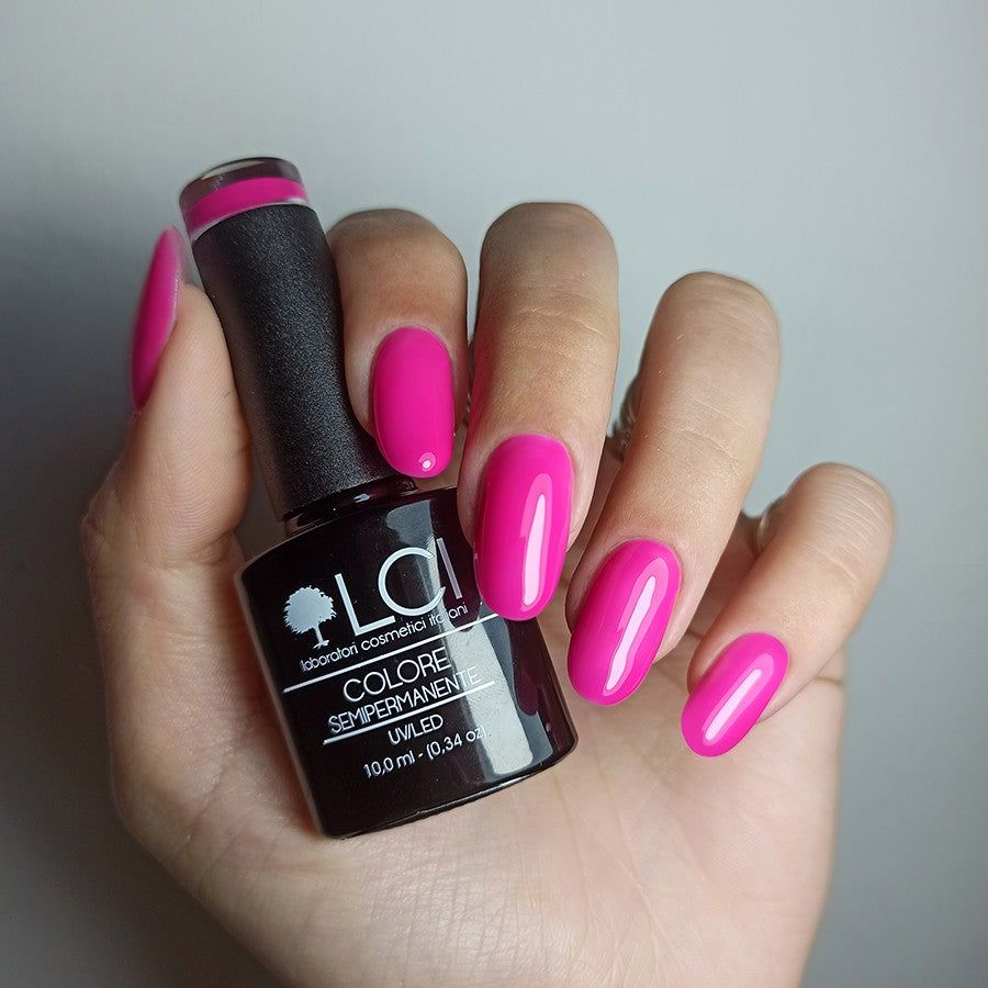 Mano che tiene flacone di smalto e unghie smaltate fucsia neon. Flacone nero con testo bianco. Unghie ovali.