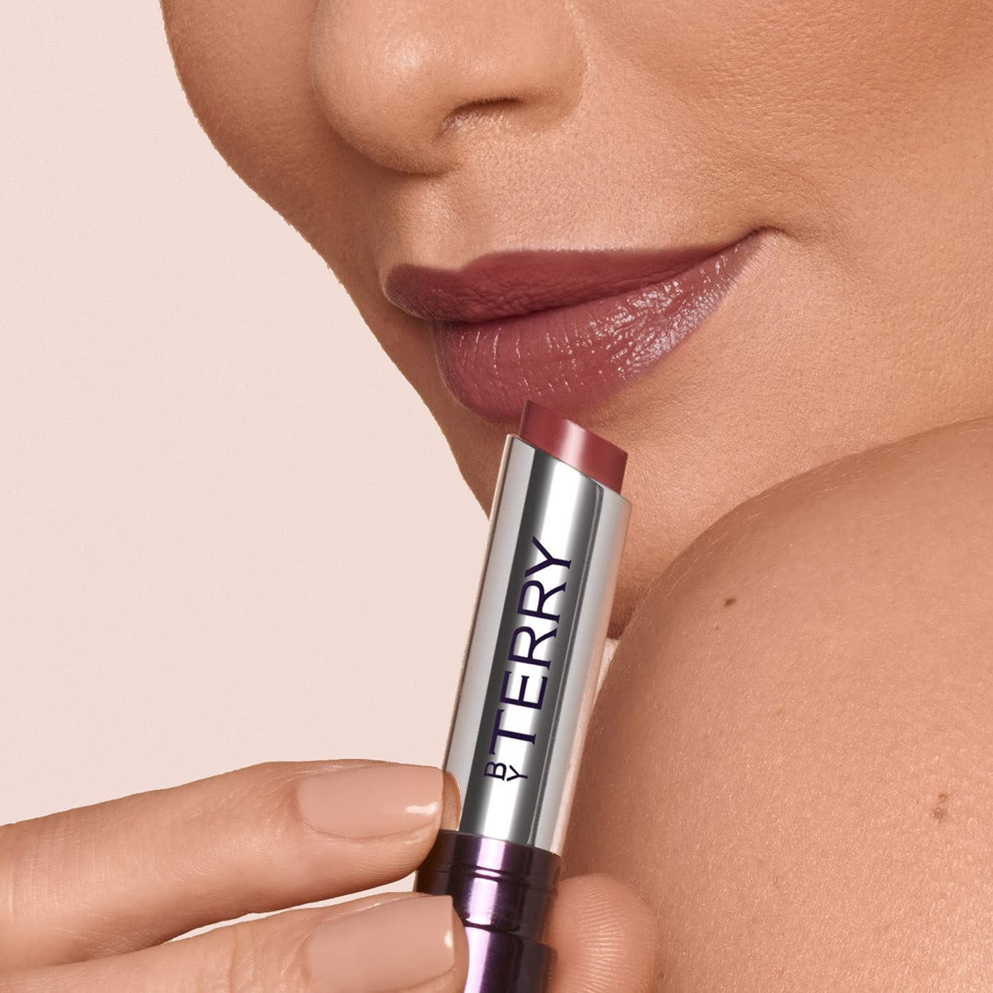 Labbra che tengono un rossetto. Tubo argento con "BY TERRY". Rossetto in uso.