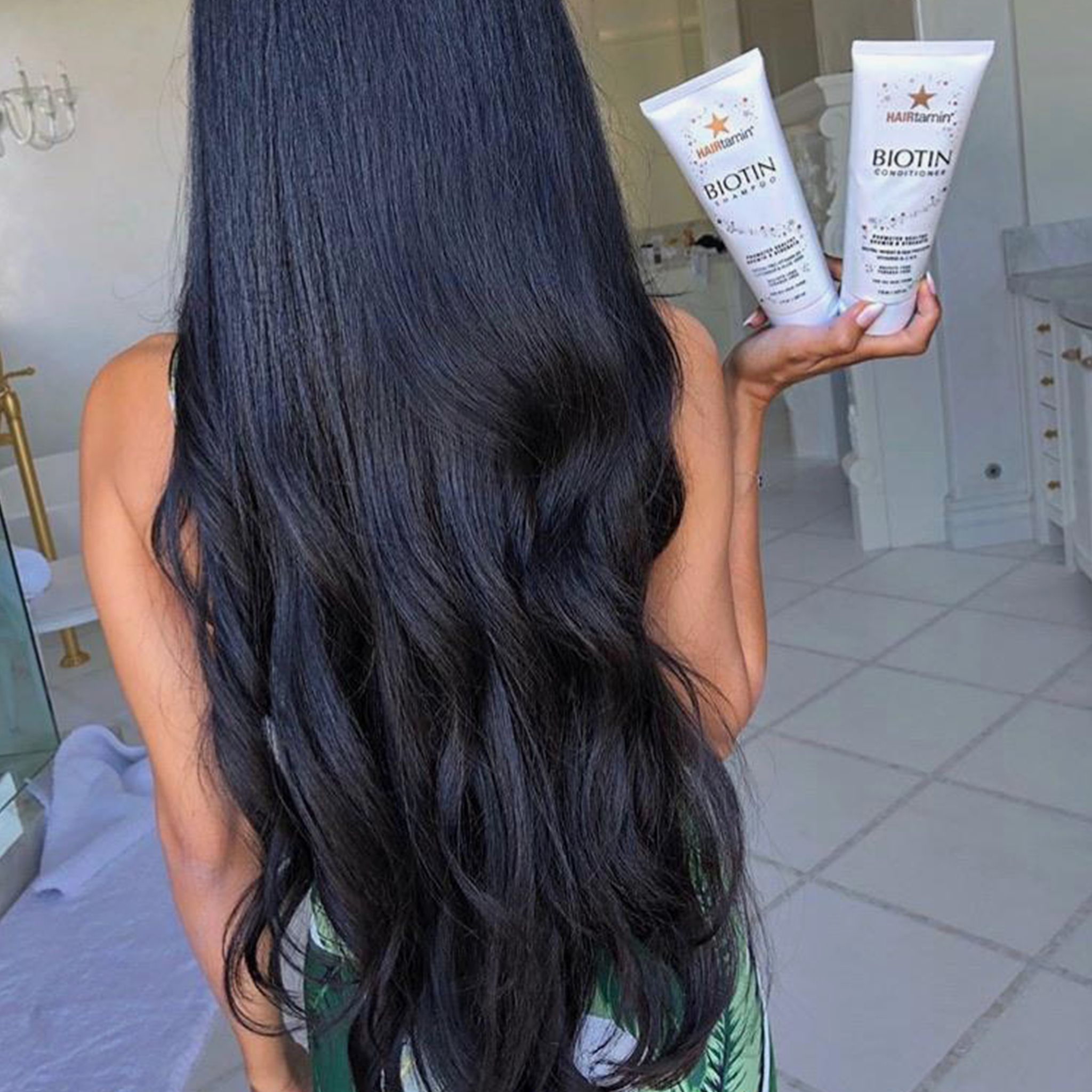Due tubi bianchi "HAIRtamin Biotin Shampoo" in mano. Capelli neri e lisci in primo piano.