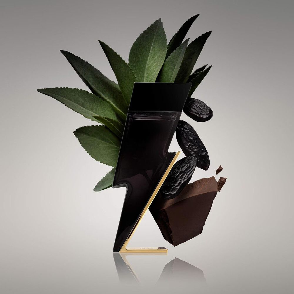 Flacone nero a forma di fulmine, base dorata, circondato da foglie, frutta e pezzo di cioccolato.