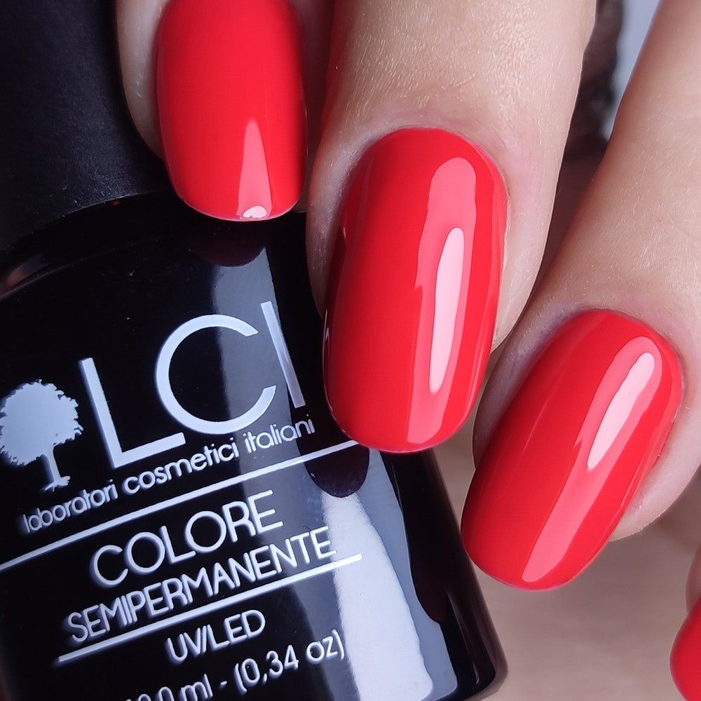 Unghie rosse lucide. Flacone di smalto nero con logo e testo bianchi. Primo piano.