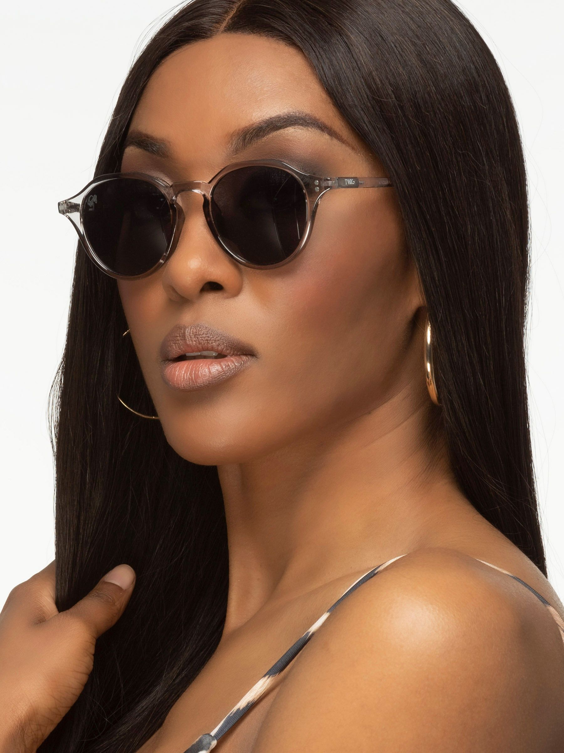 Una donna indossa occhiali da sole grigi con lenti rotonde. Gli occhiali sono sul suo naso. Indossa orecchini a cerchio dorati.