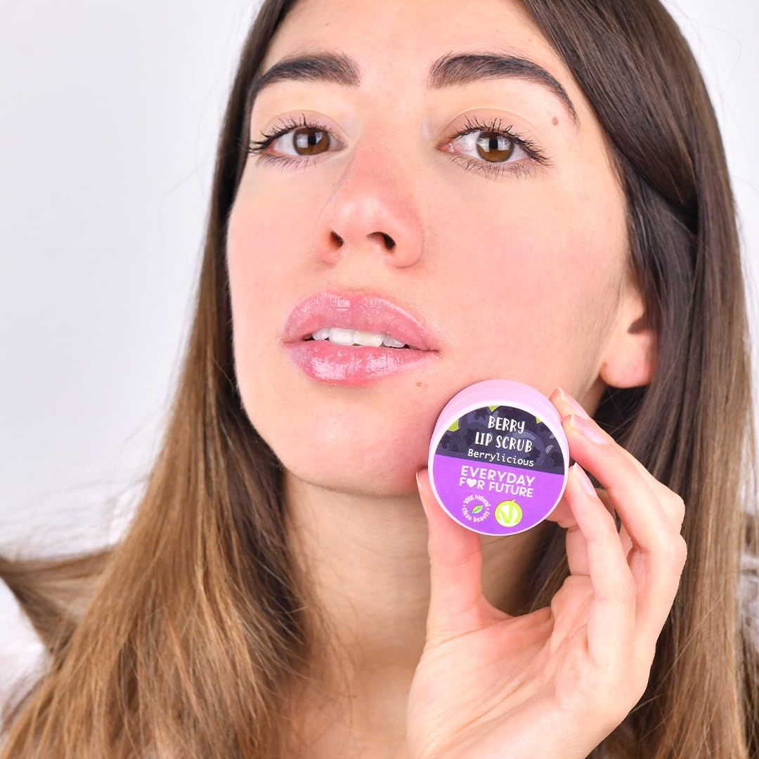 Una donna tiene uno scrub labbra rosa davanti alle sue labbra. Il nome del prodotto e il logo sono visibili sul vasetto.