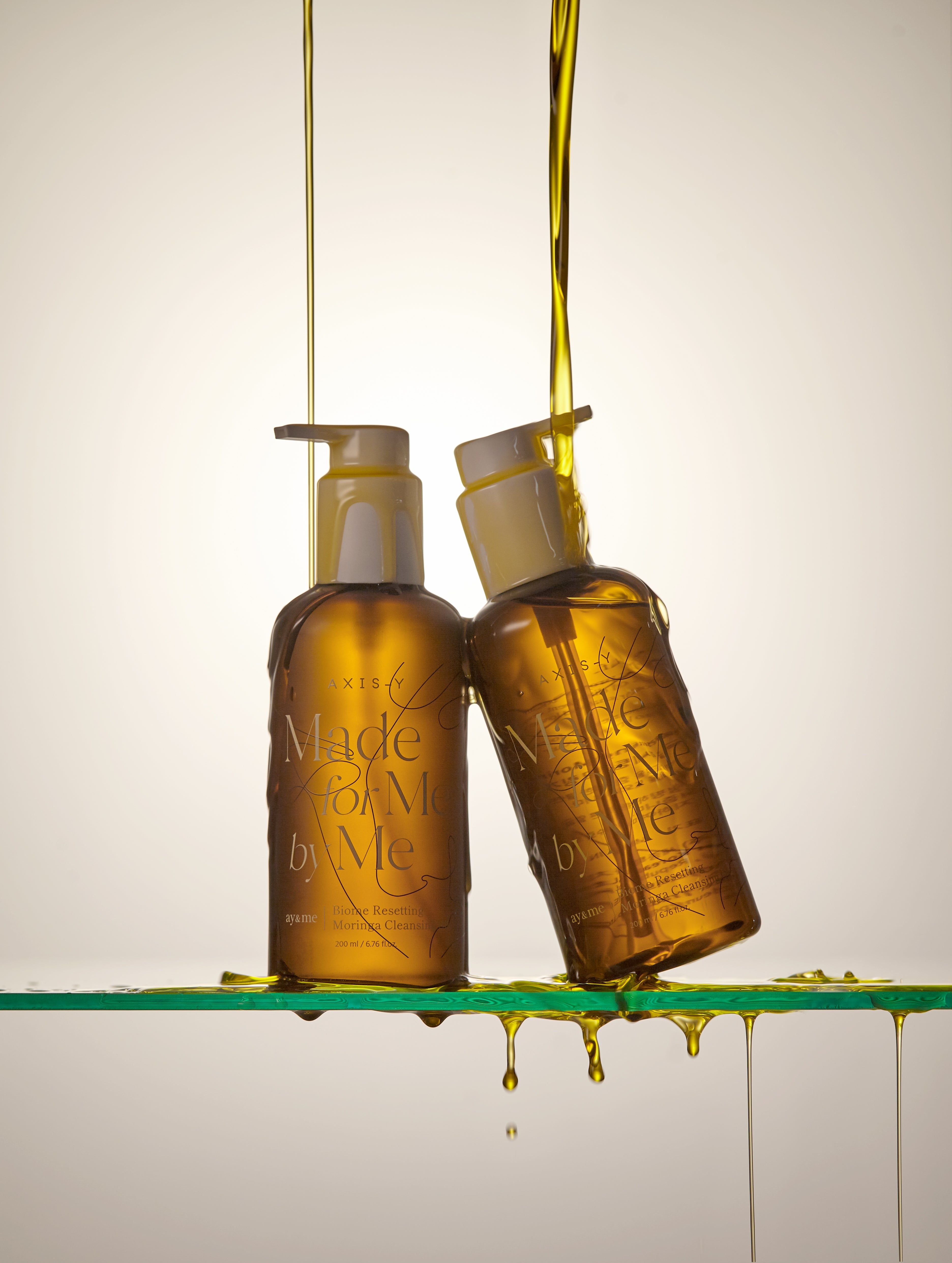 Due flaconi marroni con colata d'olio. Scritta: AXIS-Y, Made for Me by Me, Biome Resetting Moringa Cleansing Oil.