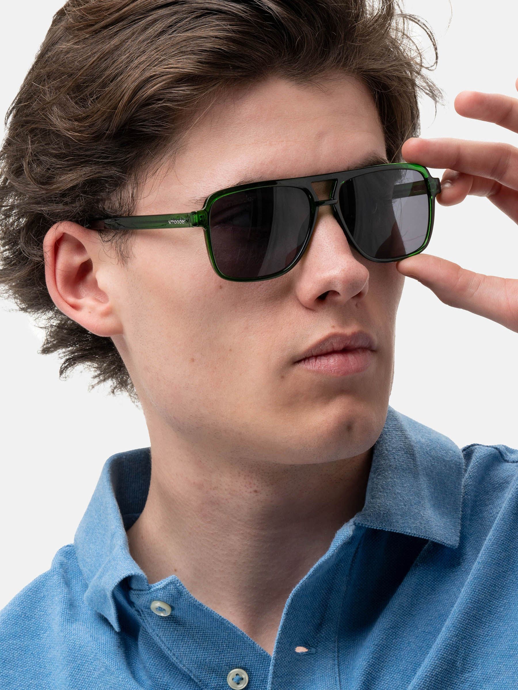Uomo tiene occhiali da sole verdi, lenti scure. Doppio ponte, marchio 'smooder' visibile.