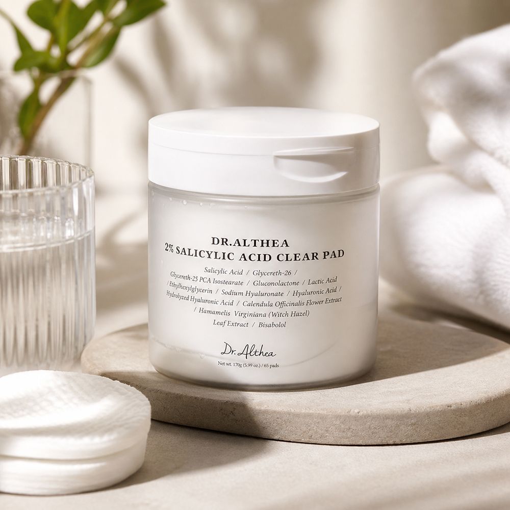 Dr. Althea 2% Salicylic Acid Clear Pad su un piedistallo di pietra, vicino a dischetti di cotone e un bicchiere.