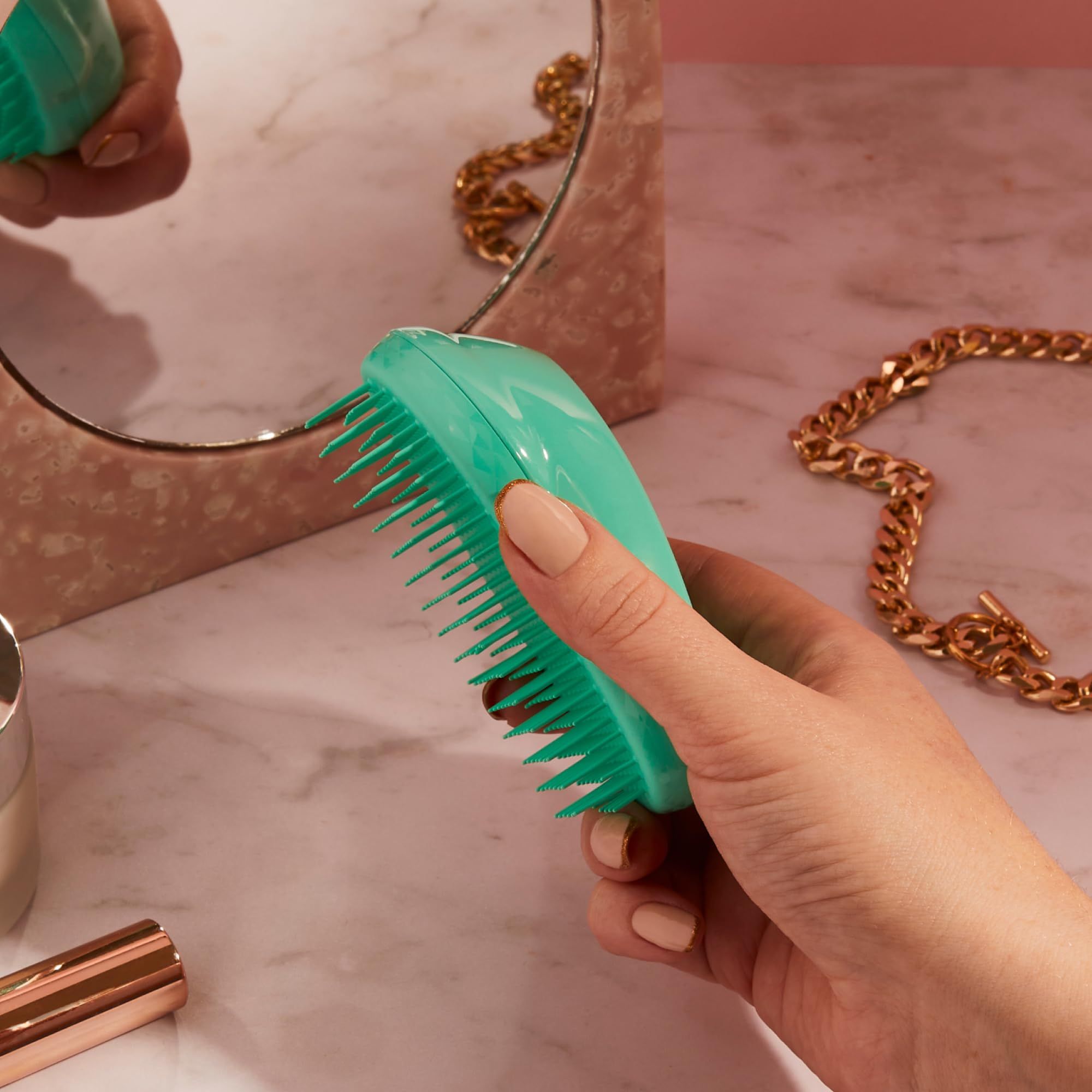 Spazzola per capelli turchese in mano. Sullo sfondo uno specchio e gioielli. La spazzola ha un'impugnatura ergonomica.