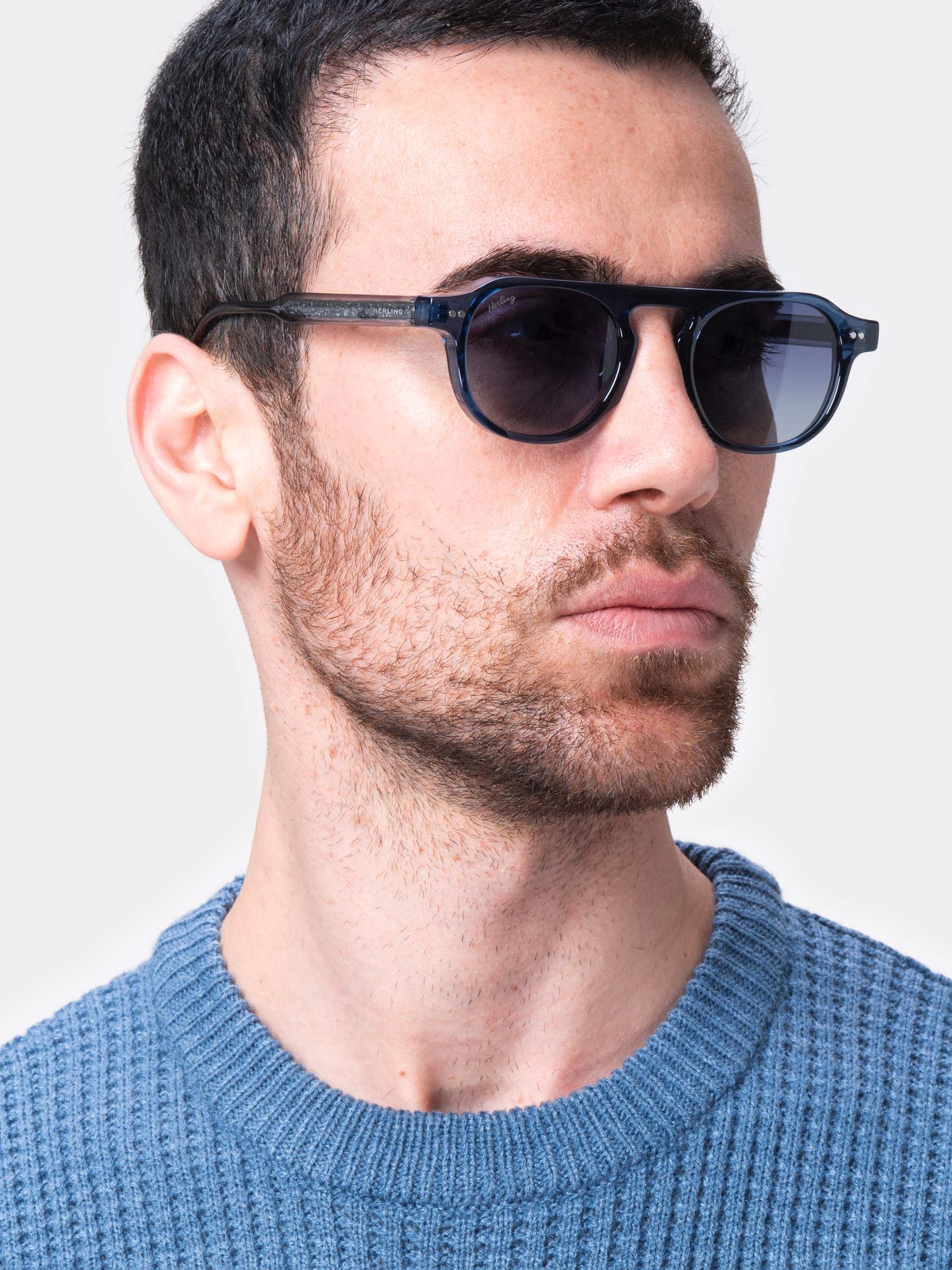 Primo piano di un uomo con occhiali da sole blu. Lenti rotonde, lenti scure, montatura blu chiaro. L'uomo indossa un maglione blu.