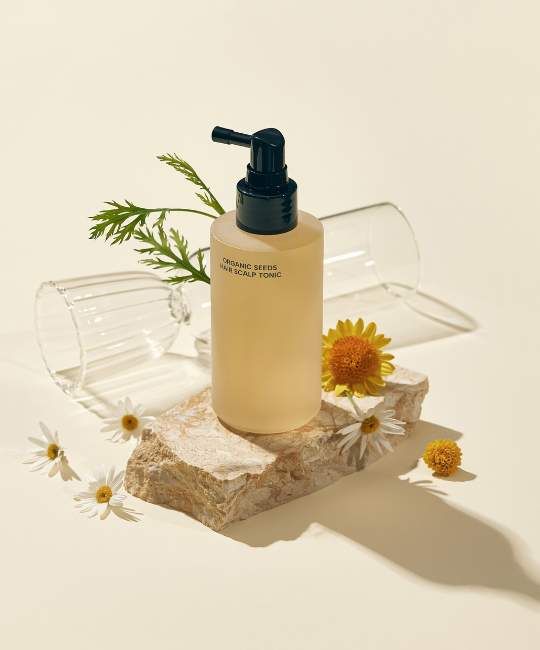 Flacone beige con erogatore nero su pietra. Scritta: ORGANIC SEEDS HAIR SCALP TONIC. Decorazioni floreali e vetro.
