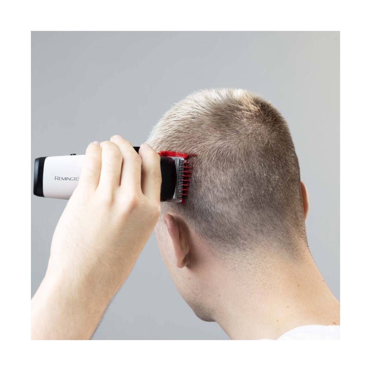 Uomo che si taglia i capelli con un tagliacapelli. Corpo bianco, pettine rosso. Sfondo grigio.