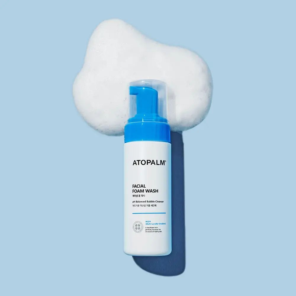 Flacone bianco-blu ATOPALM FACIAL FOAM WASH, circondato da schiuma su sfondo blu.