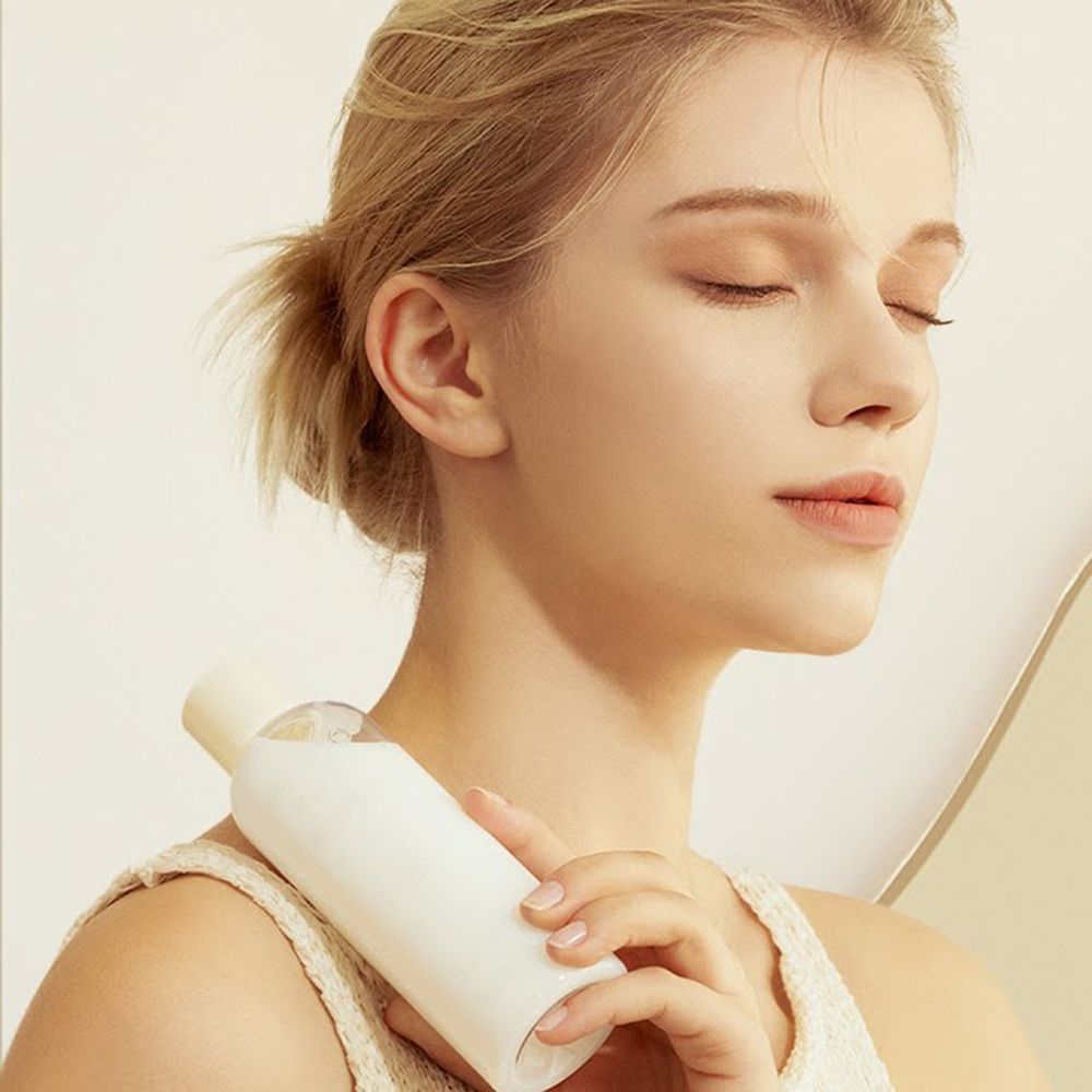 Donna tiene in mano un flacone bianco. Si tocca il viso. Lo sfondo è beige chiaro. La donna ha i capelli biondi.