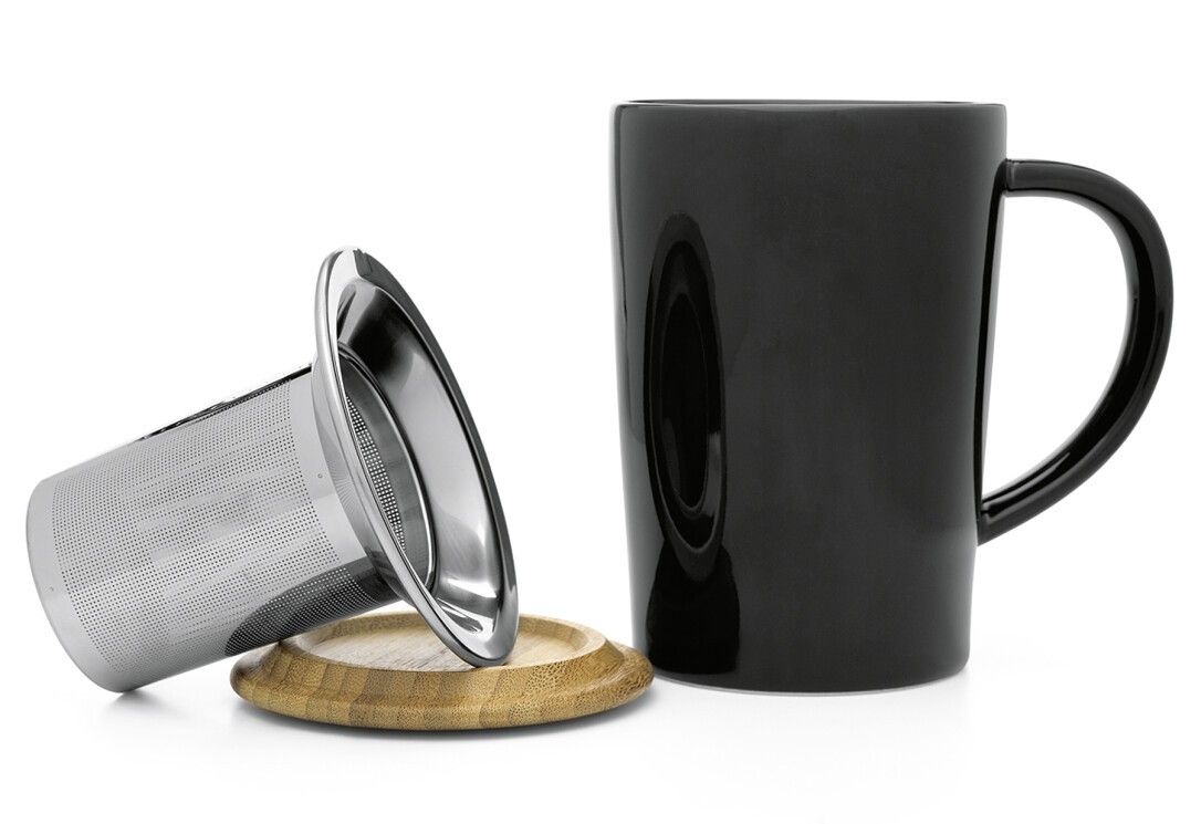 Tazza nera con manico, accanto a un colino da tè e un coperchio in legno. Su sfondo bianco.