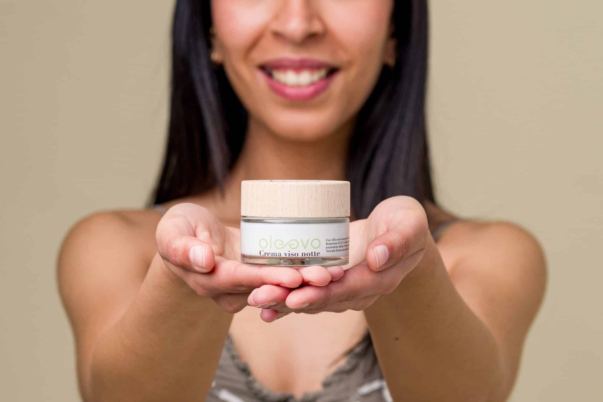 Donna tiene vasetto di crema con coperchio in legno. Scritta: Oleevo Crema viso notte. Pelle chiara, sorridente.