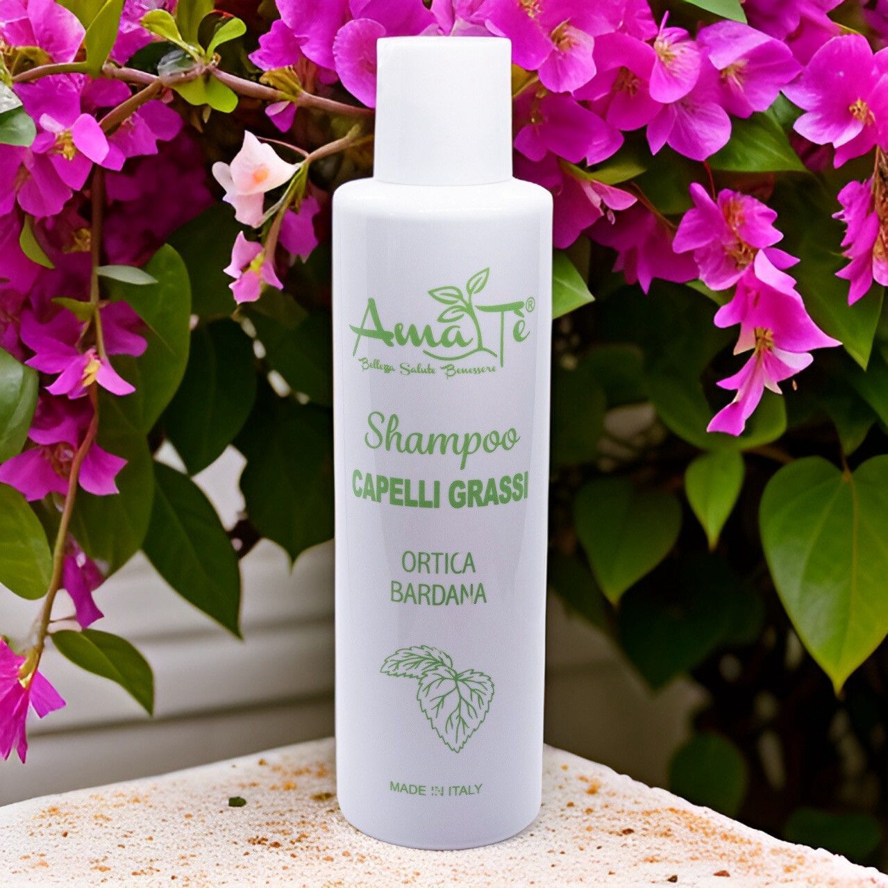 Flacone bianco con etichetta verde. Testo: Amatè Shampoo Capelli Grassi Ortica Bardana. Sfondo: fiori.