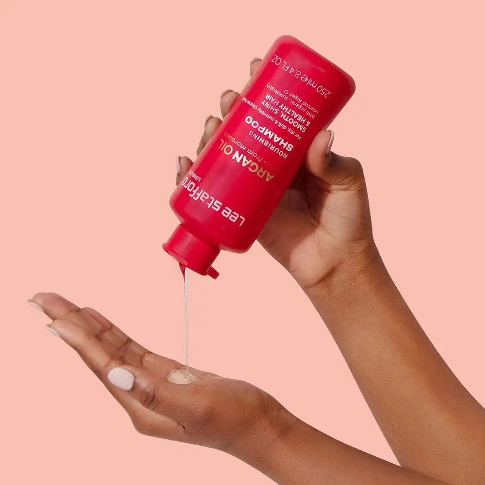 Flacone di shampoo rosso versato sulla mano. Scritta: Argan Oil, Shampoo. Mani che tengono il flacone e ricevono il prodotto.