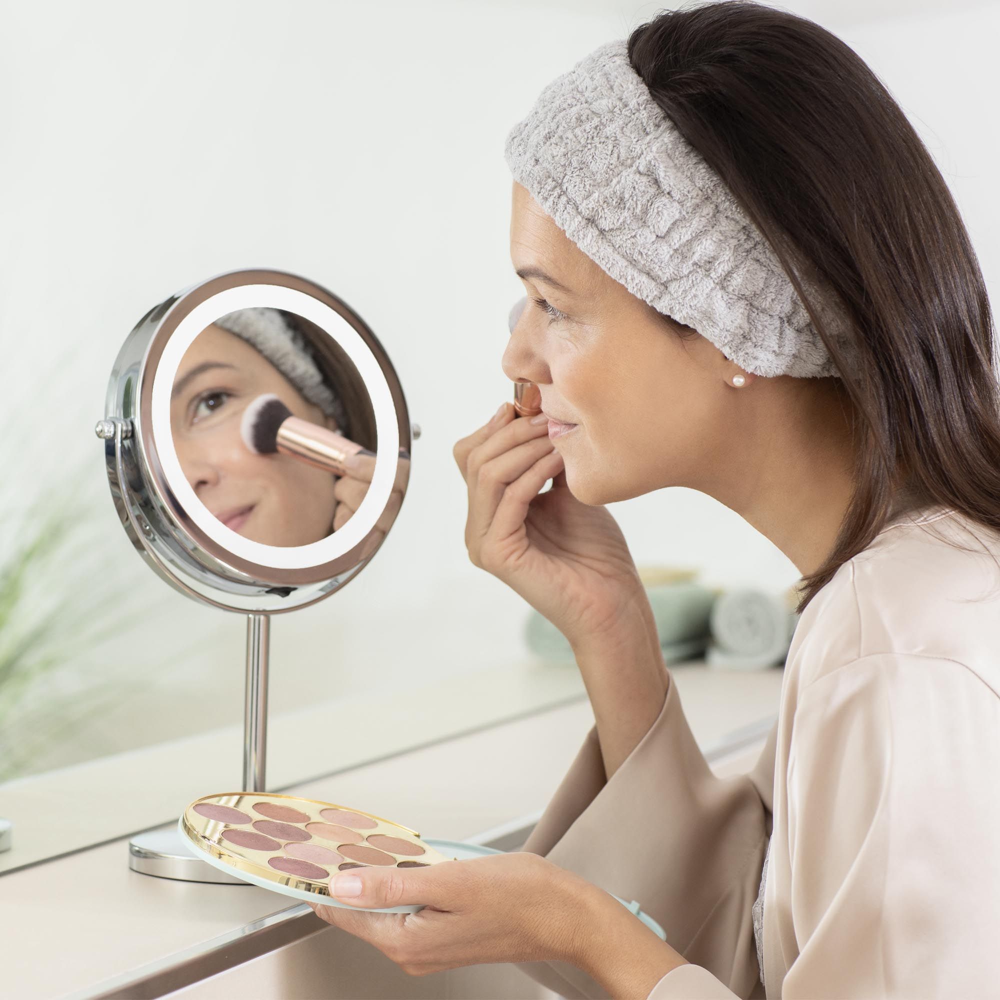 Una donna usa uno specchio cosmetico con illuminazione a LED. Indossa una fascia e tiene un pennello per il trucco.