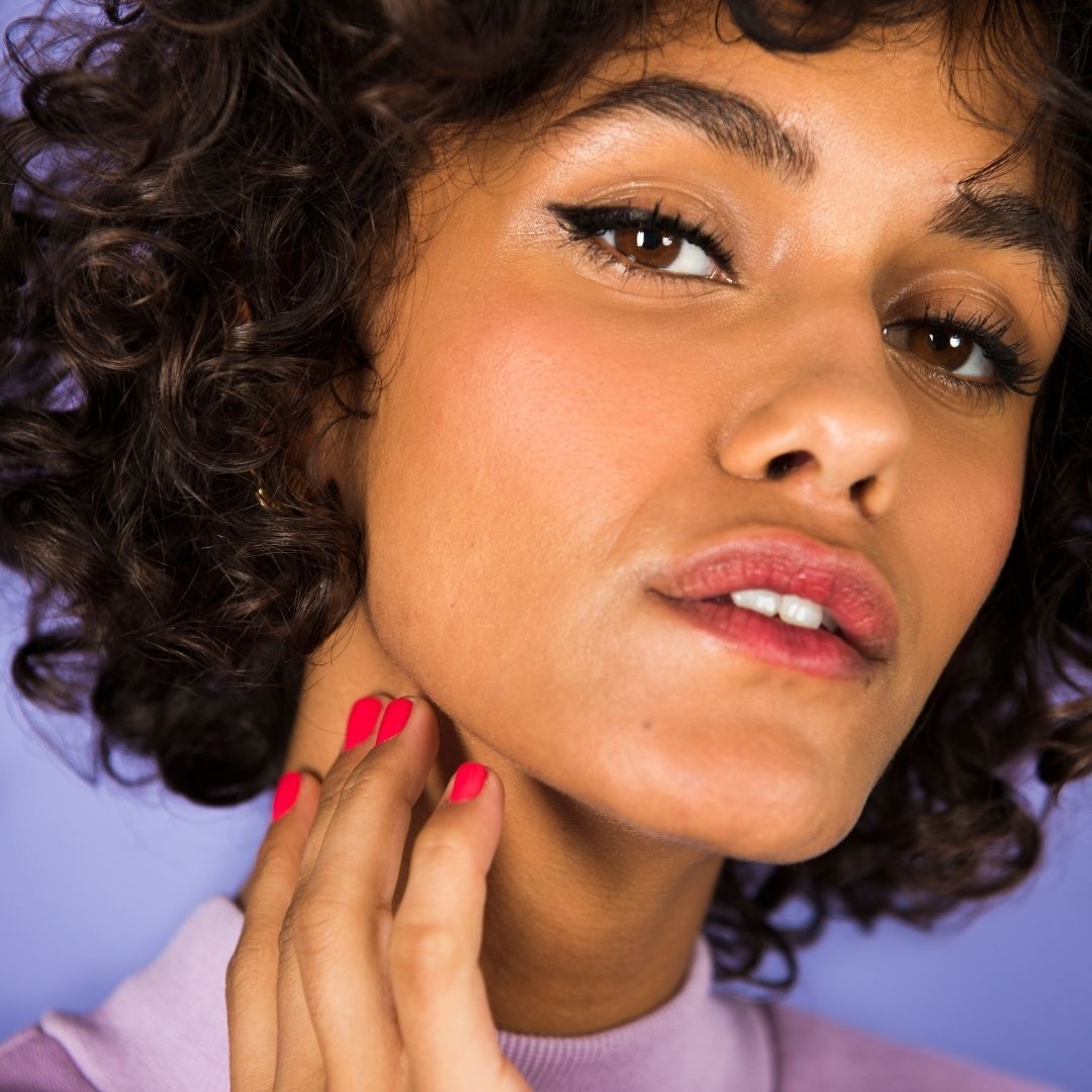 Primo piano di una donna truccata. Si tocca il collo. Visibili rossetto e smalto per unghie.