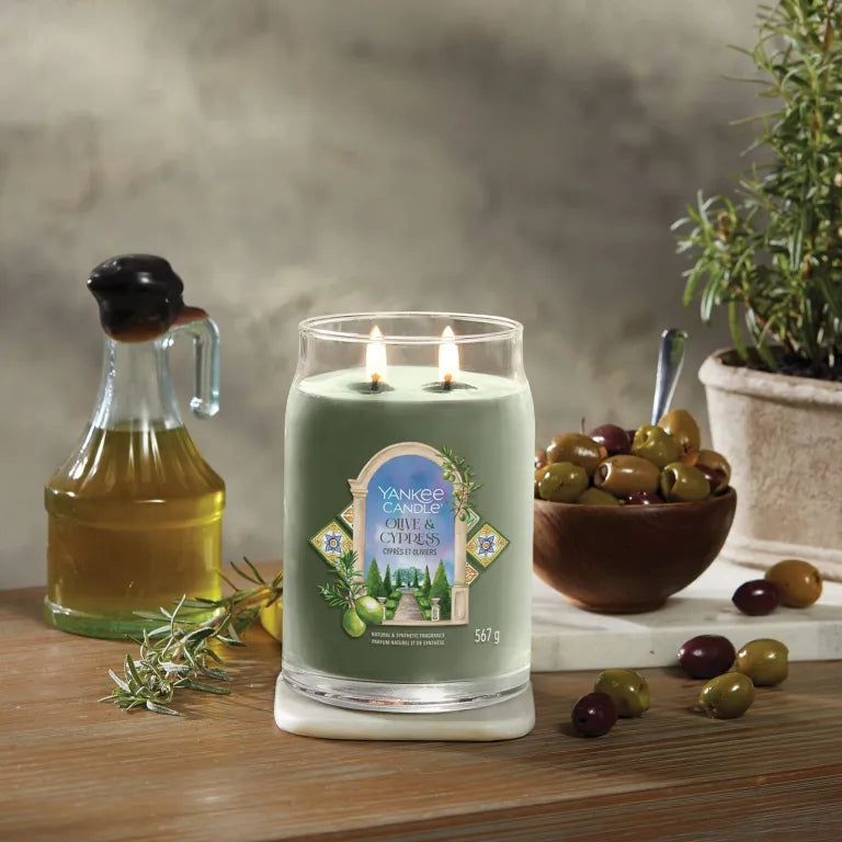 Candela Yankee Candle Olive & Cypress a due stoppini. Olive, bottiglia d'olio, rosmarino e vaso su superficie in legno.