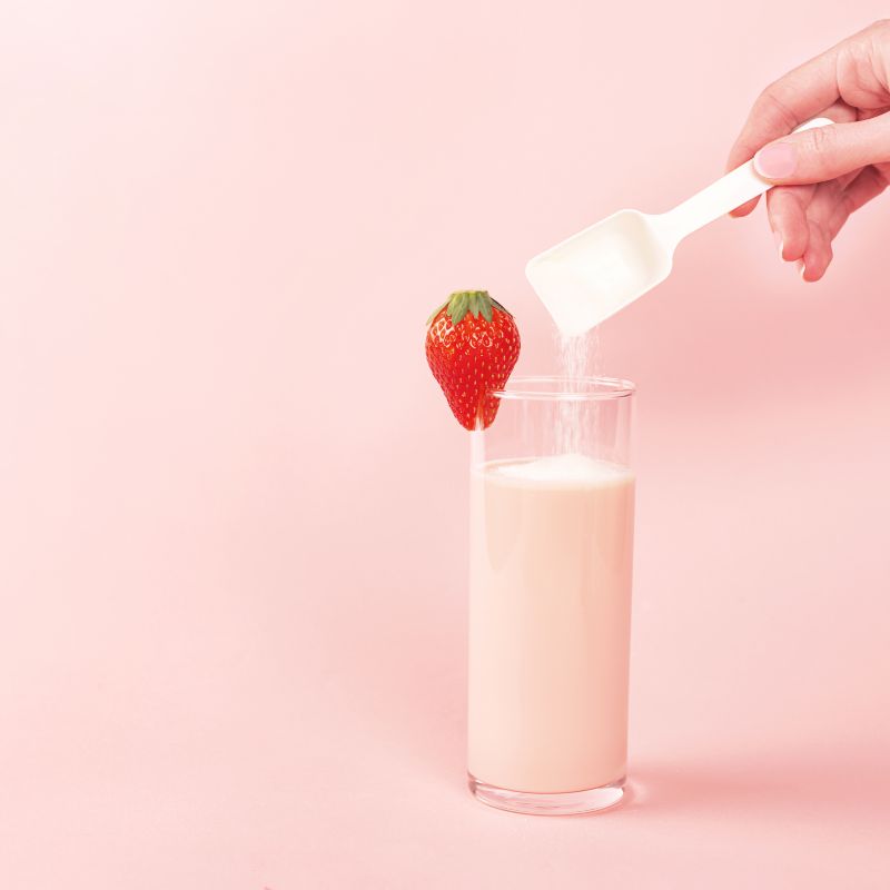 Bicchiere con liquido rosa, fragola, cucchiaio con polvere, mano. Sfondo rosa.