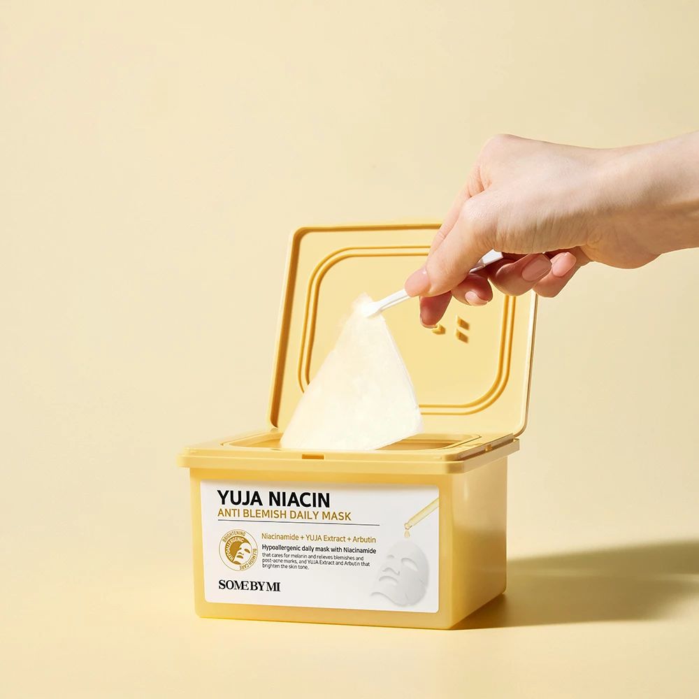 Confezione di maschere gialla aperta. Mano che estrae una maschera facciale. Scritta: Yuja Niacin Anti Blemish Daily Mask.