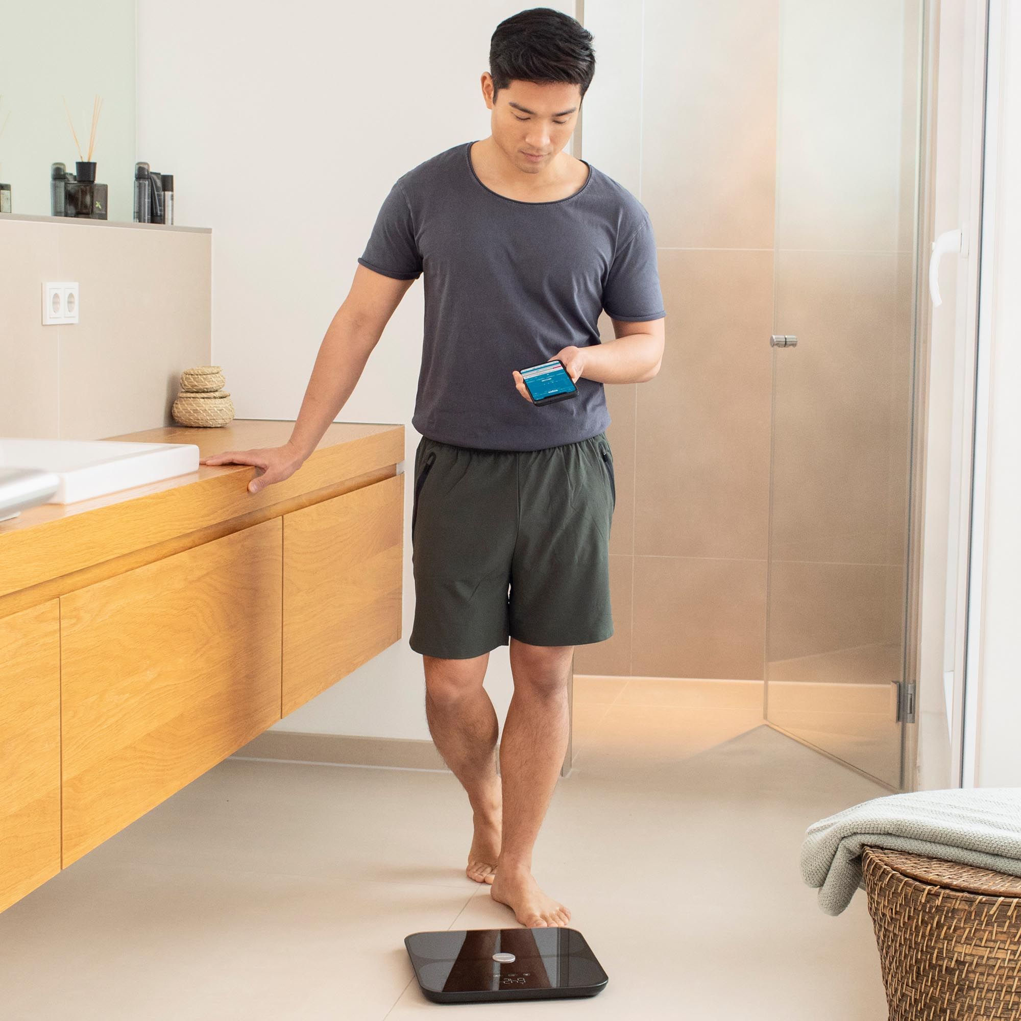 Uomo in piedi su una bilancia per l'analisi corporea nera in bagno. Tiene in mano uno smartphone.