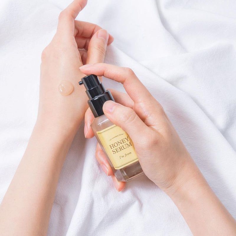 Mani che tengono un flacone di siero. Honey Serum, I'm from. Etichetta gialla, tappo nero. Siero applicato sulla mano.