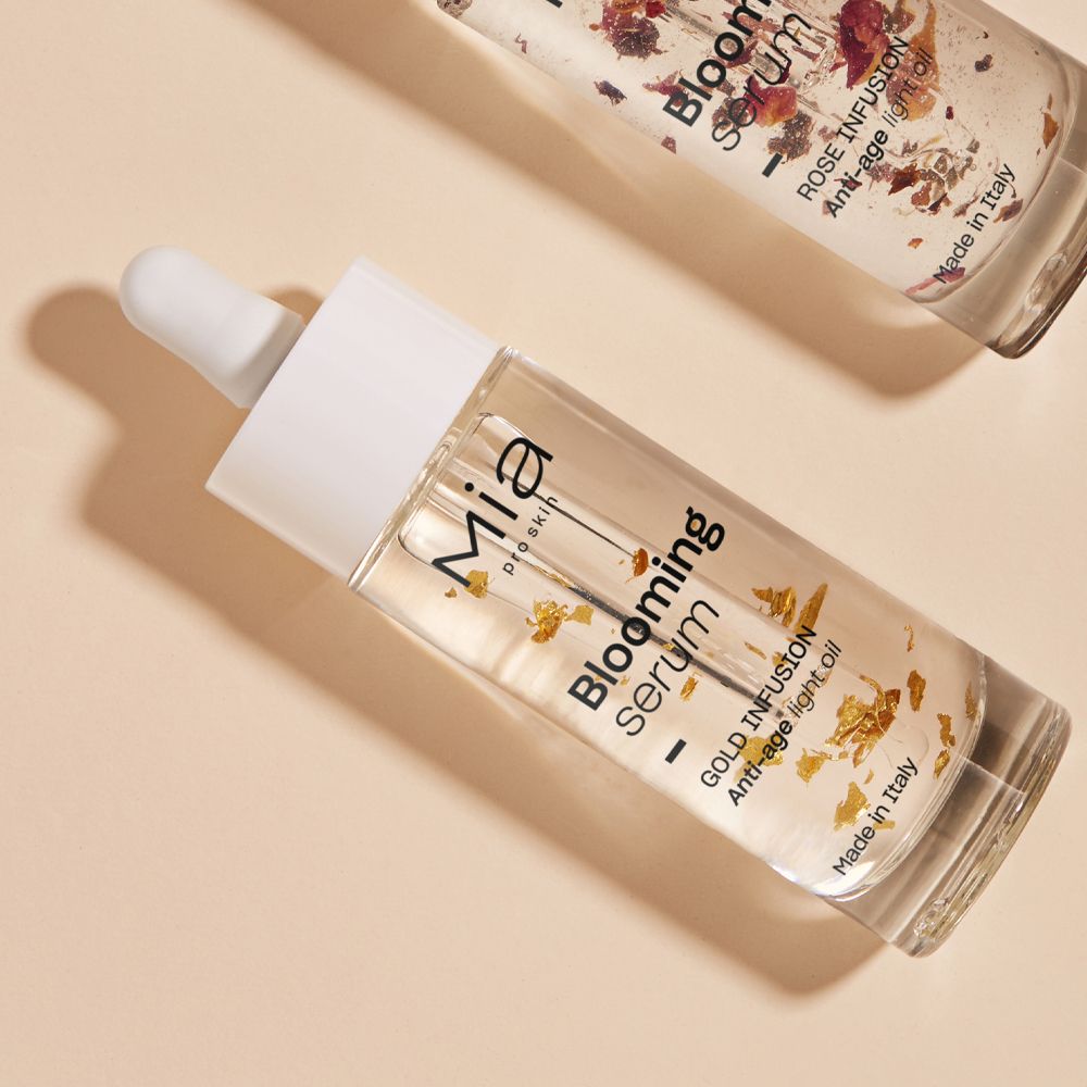Due flaconi di siero. Siero trasparente con particelle d'oro. Scritte: Mia, Blooming Serum, Gold Infusion, Anti-Age Light Oil.