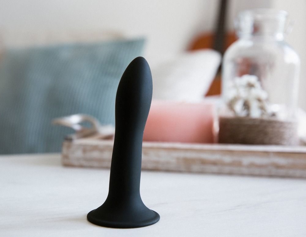 Dildo nero su superficie bianca. Base larga, forma curva. Sullo sfondo: decorazioni.