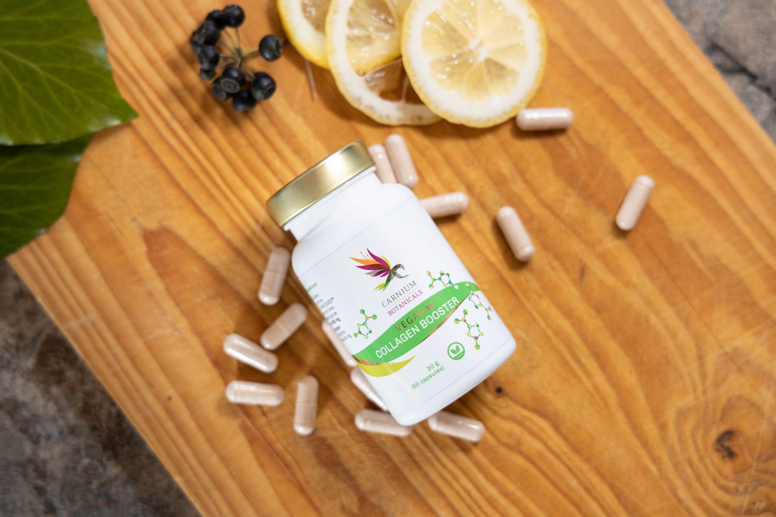Flacone e capsule su tagliere in legno. Scritta: Carnium Botanicals Vegan Collagen Booster. Fette di limone e bacche.