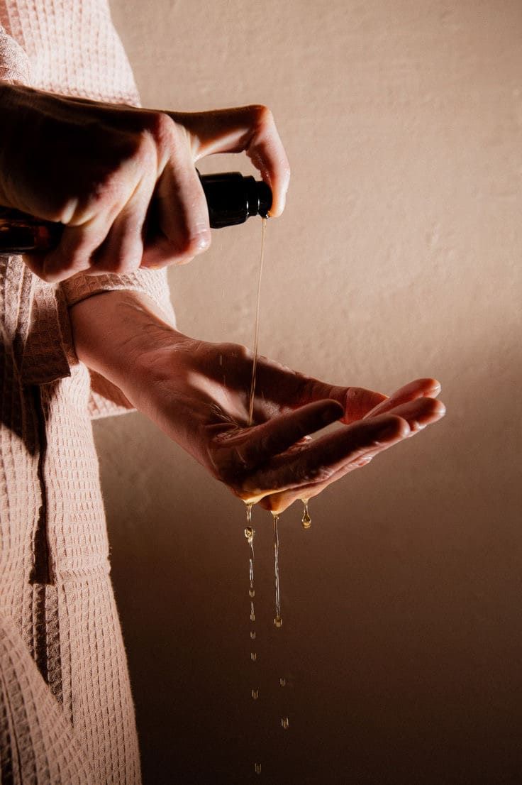 Mani che tengono un flacone nero. L'olio viene versato sul palmo. Sfondo: beige.