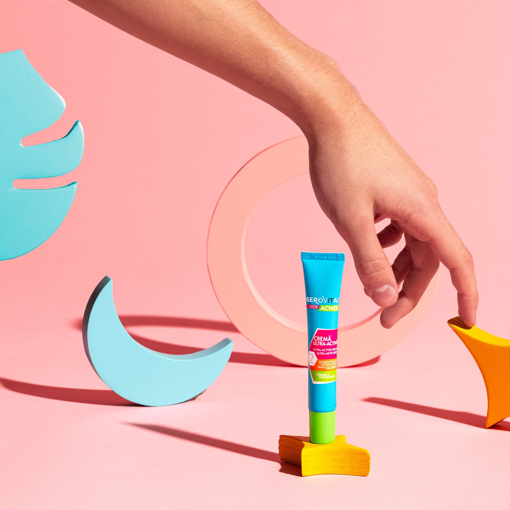 Tubo turchese e verde su piedistallo giallo, tenuto da una mano. Sfondo rosa con forme in legno. Scritta: Stop Acnee.