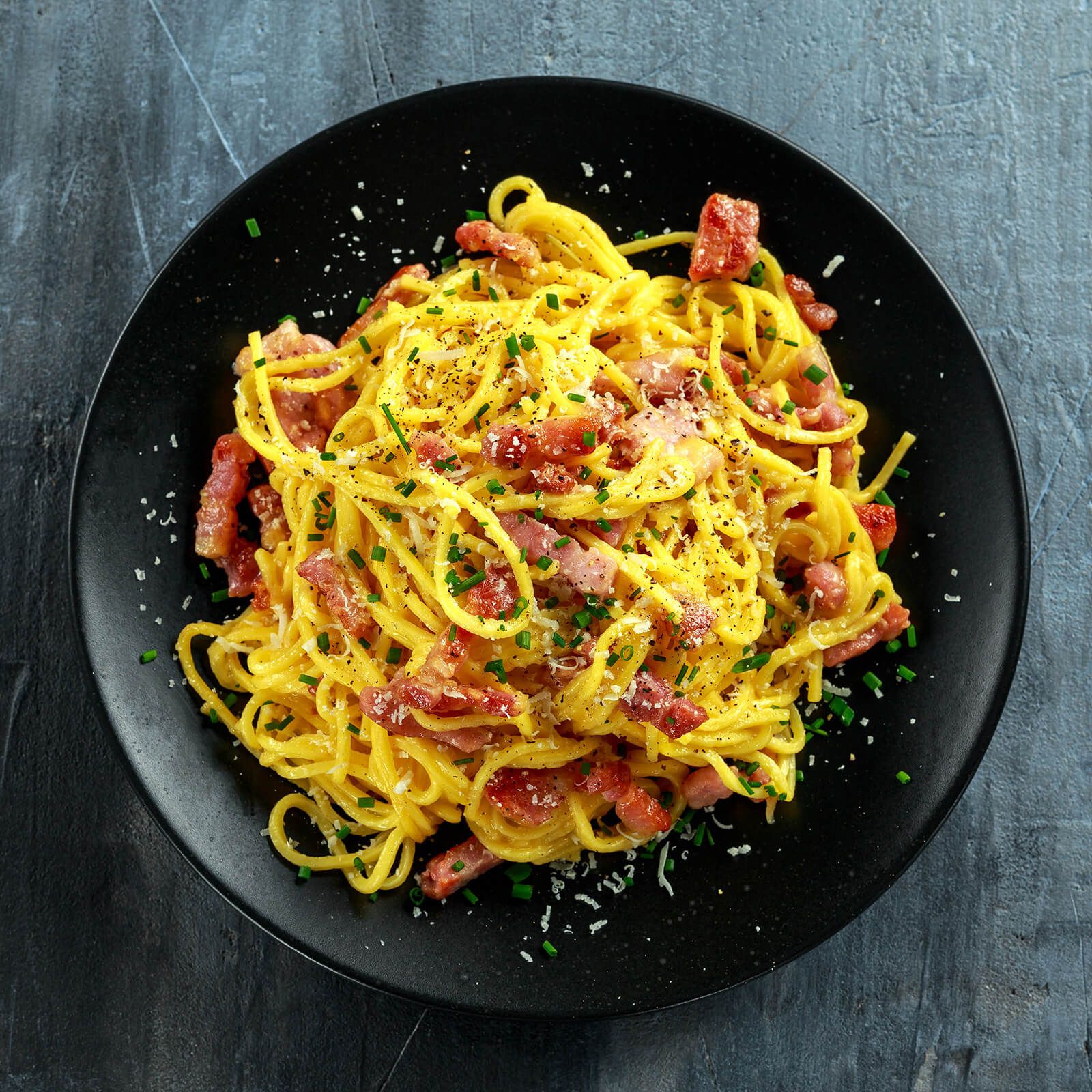 Piatto nero di spaghetti alla carbonara con pancetta, uova e formaggio. Erba cipollina.