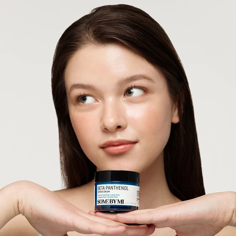 Una donna tiene in mano un barattolo di crema blu con coperchio nero. Sul barattolo si legge "BETA PANTHENOL" e "SOME BY MI".