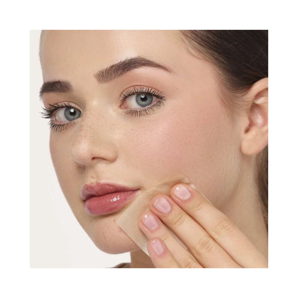 Donna con incarnato chiaro usa una carta assorbente quadrata sulla pelle. La carta tocca la guancia.